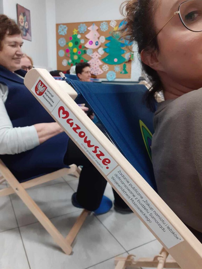 widok na brzeg drewnianego leżaka na którym jest nalepka z napisem: Zadanie publiczne "Zakup namiotu i leżaków" dofinansowane z budżetu Samorządu Województwa Mazowieckiego. Na leżaku siedzi kobieta. W tle kilka innych pań siedzi także na leżakach. Zakupiliśmy namiot oraz 20 leżaków dzięki dofinansowaniu z Urzędu Marszałkowskiego Województwa Mazowieckiego w Warszawie.