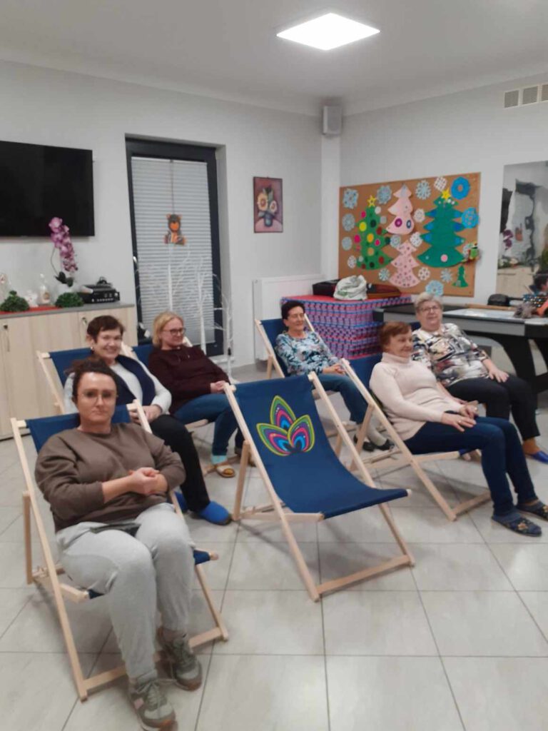 Grupa 6 pań siedzących na drewnianych leżakach w świetlicy wiejskiej. Jeden leżak jest pusty. Widać na nim logo KGW Pawie Oczko. Zakupiliśmy namiot oraz 20 leżaków dzięki dofinansowaniu z Urzędu Marszałkowskiego Województwa Mazowieckiego w Warszawie.