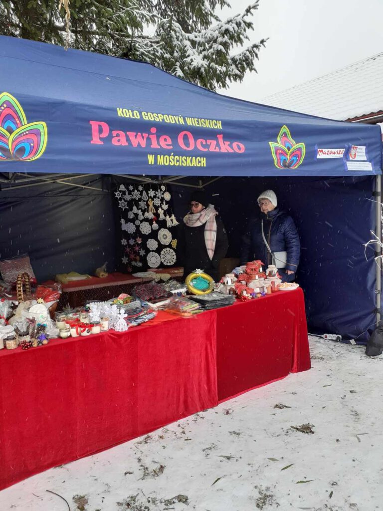 Widok na stoisko straganowe z wyrobami ręcznymi Koła Gospodyń Wiejskich. na stole z czerwonym materiałem. Dwie panie stoją pod granatowym namiotem z logo KGW Pawie Oczko w Mościskach oraz podpisem z nazwą koła. Zakupiliśmy namiot oraz 20 leżaków dzięki dofinansowaniu z Urzędu Marszałkowskiego Województwa Mazowieckiego w Warszawie.